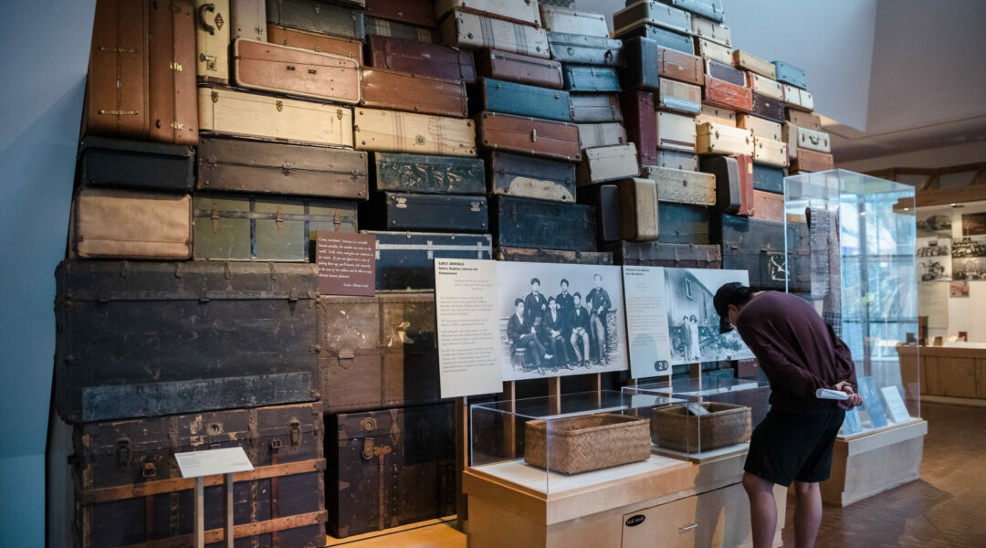 JANM common ground suitcases A visitor leans over a display case to look at objects in an exhibition gallery. Behind the case is a wall of suitcases stacked on top of each other.