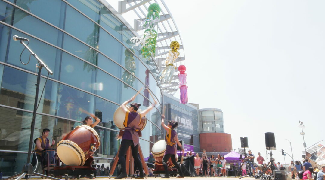 2023.08.12-natsumatsuri-taiko-proj-janm-cooper-cuya-3 A taiko group performs outside of the Japanese American National Museum. The museum building is behind them in the background with colorful streamers hanging from the top of the building. A large crowd is surrounding the taiko performers.